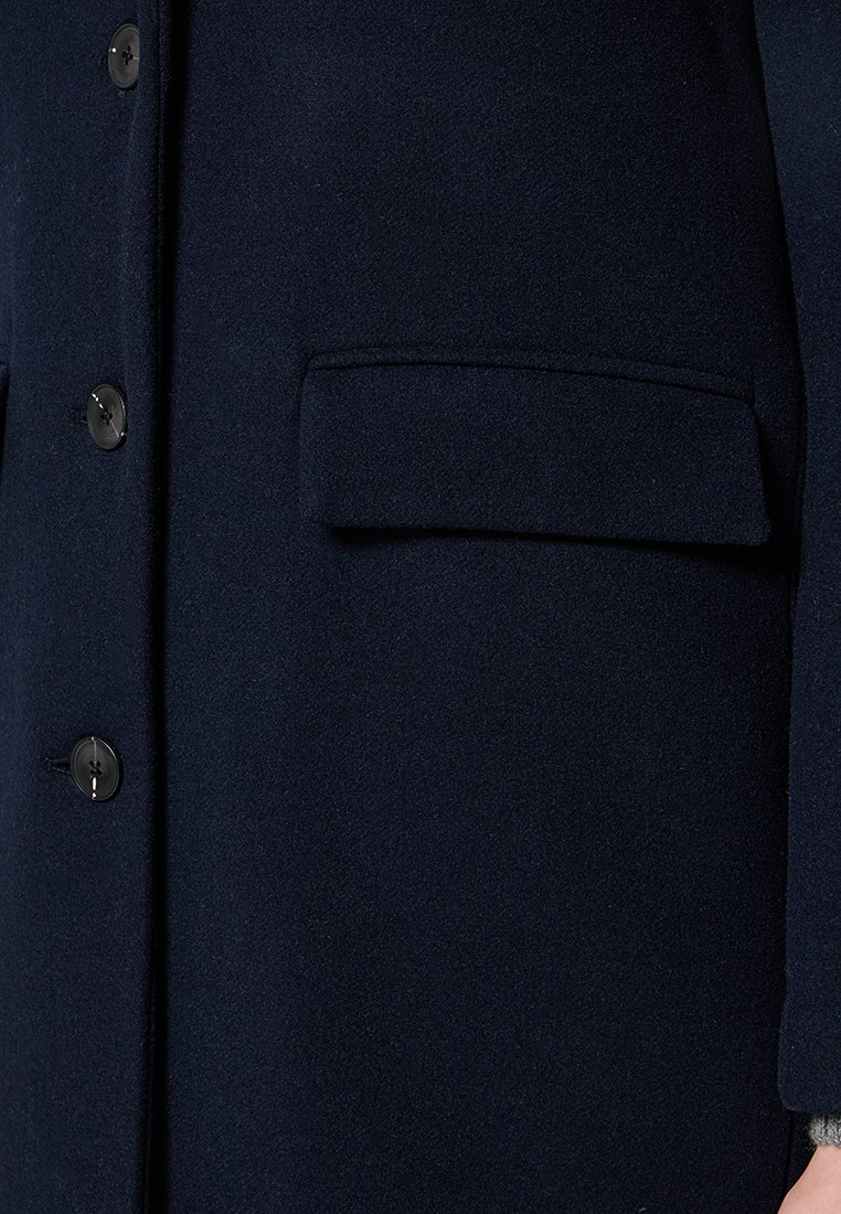 Marineblauer Wollmantel mit glatter Textur, ausgestattet mit einer Brusttasche, schwarzen Knöpfen und einem klassisch-taillierten Design. Nahaufnahme der vorderen Details.