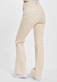 Bakgård av en person iført lysebeige slengbukser og hvite sneakers, stående mot en ensfarget hvit bakgrunn.