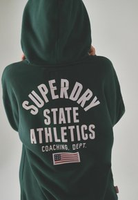 Felpa con cappuccio verde scuro con scritta bianca: "SUPERDRY STATE ATHLETICS COACHING. DEPT." Include un piccolo dettaglio con la bandiera americana in basso.
