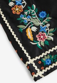 Tissu noir avec des motifs floraux brodés colorés en bleu, rose, orange, vert et blanc, bordé d'un galon ondulé crème et de petits points orange.
