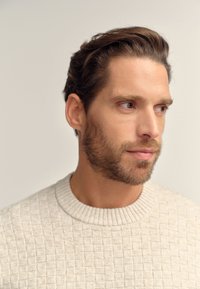 Ljust beige stickad tröja med ett strukturerat rutmönster, ribbad halsringning och medelviktigt tyg. Modellen har kort skägg och rufsigt hår.