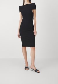 Abito midi nero off-the-shoulder con texture a costine, vestibilità aderente e tessuto morbido. Abbinato a mule nere con la punta aperta.