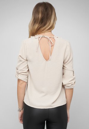 Frau mit langen Haaren trägt eine hellbeige Bluse mit hochgekrempelten Ärmeln und einem gebundenen Schlüsselloch am Rücken, kombiniert mit einer schwarzen Hose.