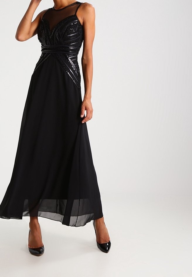 Robe de soirée noire sans manches avec corsage à sequins et encolure transparente, portée avec des talons noirs vernis à bout pointu sur un fond neutre.