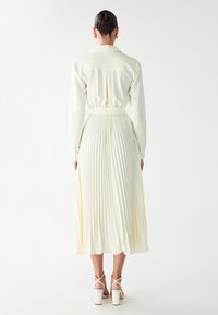 Cremefarbenes, in Falten gelegtes Kleid mit langen Ärmeln, einem Kragen und einer taillierten Taille. Hat eine weiche Textur und fällt unter das Knie.