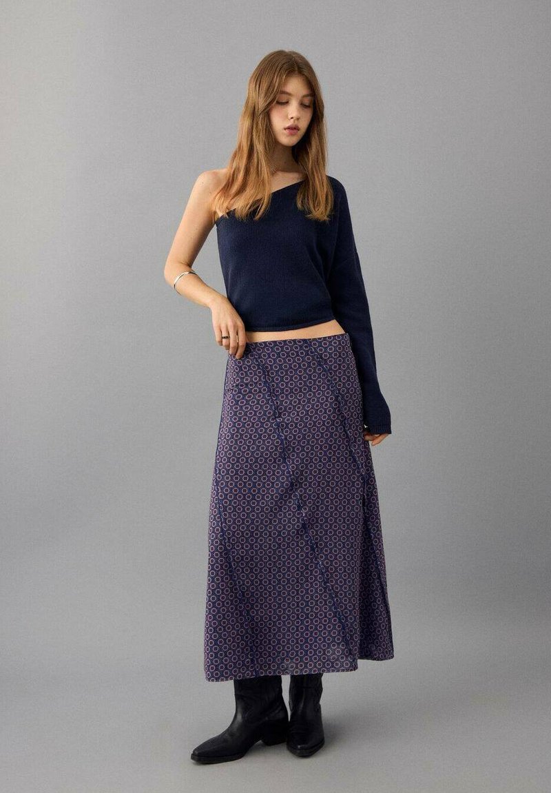 Navy one-shoulder sweater, paired with a long, patterned skirt featuring circles in red and blue. Black ankle boots complete the look.