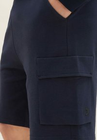 Námořnické modré šortky s velkou postranní kapsou a malým černým logem poblíž spodní části, nošené s rukou v kapsičce na boku.