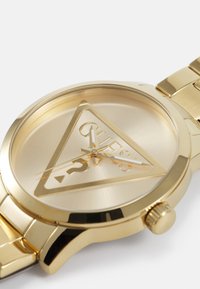 Reloj metálico dorado con una esfera triangular, que presenta un logotipo, marcas horarias blancas y tres manecillas. La correa está compuesta por segmentos metálicos enlazados.