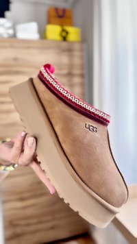 Šviesiai rudos sukurto zomšos slip-on tipo bateliai su storu smėlio spalvos padu, turintys raudonai baltą raštuotą apdailą aplink kulkšnis ir iškilų „UGG“ logotipą.
