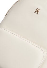 Μπεζ σακίδιο με λεία υφή, διακοσμημένο με χρυσό λογότυπο και τσέπη με φερμουάρ. Απλός σχεδιασμός με απαλά άκρα και ελάχιστες λεπτομέρειες.