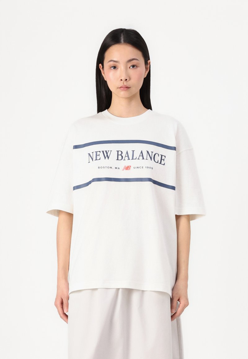 Balta kokvilnas T-kreklu ar īsām piedurknēm, ar tumši zilu grafiku "NEW BALANCE" un tekstu no Bostona, MA. Brīva piegriezuma un gluda tekstūra.
