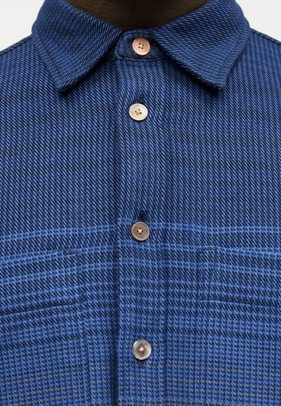 Μπλε πουκάμισο με υφή, με τέσσερα κουμπιά μπροστά και μυτερό γιακά, που δείχνει λεπτομερή ύφανση υφάσματος και ραφές.