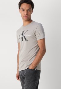 Pelēka kokvilnas t-kreklu ar lielu melnbalto "Calvin Klein Jeans" logotipu uz priekšpuses. Īsas piedurknes un klasisks apaļais kakla dizains.