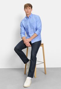 Camicia azzurro chiaro con bottoni e taschino sul petto, abbinata a jeans scuri in denim e scarpe da ginnastica bianche. Modello seduto su uno sgabello di legno.