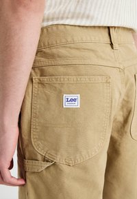 Kaki arbeidsbukser i strukturert bomullsmateriale, med en baklomme og en blå og hvit "Lee Workwear" etikett.