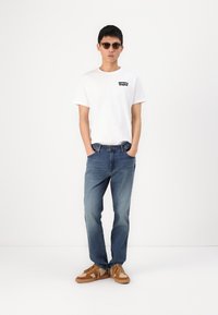 Bijela pamučna majica s crnim Levi's logotipom, plave traperice i smeđe sueđe cipele s teksturiranim akcentima. Model nosi sunčane naočale i stoji opušteno.