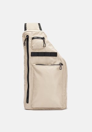 Beige crossbody skuldertaske med flere lynlåslommer og sorte stropdetaljer, vist opret mod en ensfarvet baggrund.