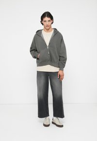 Harmaa vetoketjuhuppari, jossa on etutasku, kerroksittain vaaleancremepaidan päällä, yhdistettynä leveälahkeisiin harmaisiin farkkuihin ja vaaleisiin tennareihin.