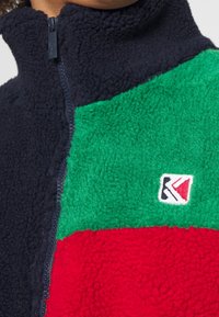 Fleecová bunda s barevnými bloky v námořnické modři, zelené a červené. Vysoký límec se zapínáním na přední zip. Vyšitá značka na pravé dolní straně.