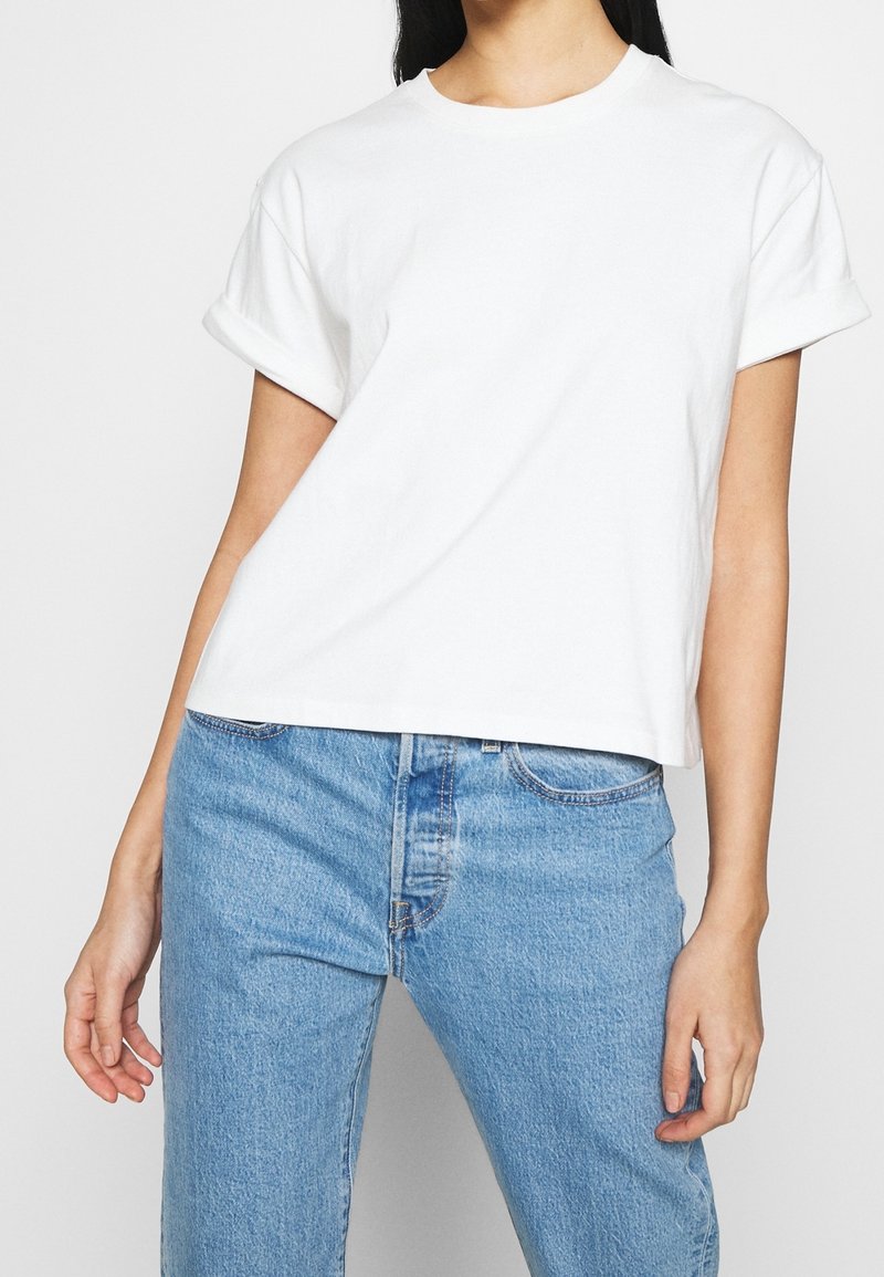 Weißes Baumwoll-T-Shirt mit umgeschlagenen kurzen Ärmeln, Rundhalsausschnitt und leicht verkürztem Saum, getragen mit hellblauen Jeans. Glatte Textur.
