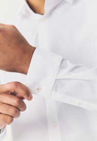 Uomo che aggiusta il polsino di una camicia bianca a manica lunga, concentrandosi sulle mani e sui dettagli della camicia.