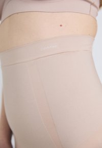 Beige shapewear met hoge taille, gemaakt van gladde, rekbare stof en een Calvin Klein-logo in de buurt van de tailleband. Platte naden en minimale textuur.