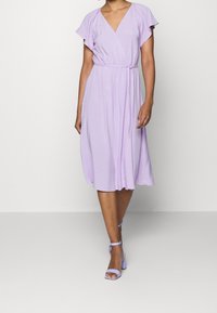 Levandulové šaty do poloviny stehna s volánkovými krátkými rukávy, elastickým pasem, hladkým materiálem a uvolněným střihem. Kombinované s levandulovými podpatky.