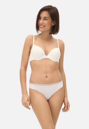 Mujer sonriente con cabello corto y castaño, vistiendo un sujetador blanco acolchado y bragas claras con estampado ligero, de pie frente a un fondo liso y claro.