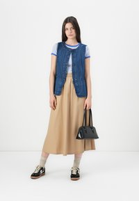Mujer joven vestida con una camiseta a rayas, chaleco de mezclilla, falda midi beige, zapatillas y sosteniendo un bolso negro frente a un fondo blanco liso.