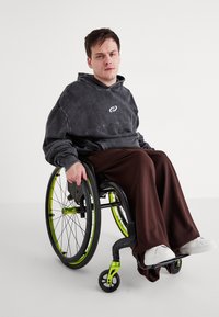 Pelēks lielgabarīta kapuce ar spirālveida logotipu, brūni plaša kāju bikses un baltas čības. Ratiņkrēsls ar melnām riteņiem un zaļiem akcentiem.