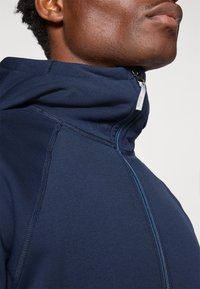 Tumedast sinise tõmbluku ja kõrge kraega lukuga jope, mis on valmistatud pehmest kangast. Omab kontrastset sinist tõmblukku ja sujuvat õlavarre disaini, mis annab voolava ilme.