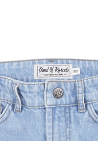 Vaaleansiniset farkkushortsit, joissa on vyötärönauha, metallinen nappi ja etiketti, jossa lukee "Raskarien bändi." Koko vaatteessa on ompelukoristeluja.