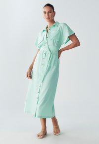 Vestito camicia verde menta con maniche corte, chiusura frontale con bottoni, tasca decorativa, vita con cintura e lunghezza midi. Notevole per la sua texture liscia.