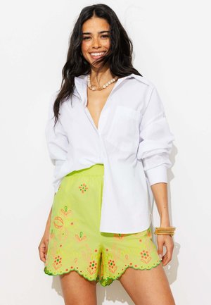 Femme souriante, portant une chemise blanche boutonnée, un short jaune brodé, un collier de perles et des bracelets empilés, debout sur un fond blanc.