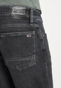 Mustat farkut, joissa on nahkapatentti vyötäröllä, johon on painettu "TOMMY JEANS". Huomaamaton ompeluseikka takataskuissa, joissa on logovarma.