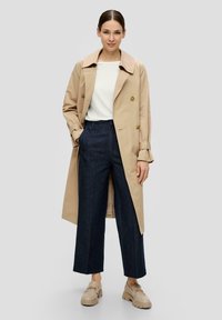 Beige langer Trenchcoat mit doppelreihigem Design und goldenen Knöpfen, kombiniert mit dunkelblauen weit geschnittenen Hosen und beigen Loafers.