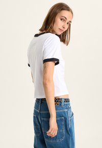 White crop t-shirt with black ribbed collar and sleeves, paired with high-waisted blue denim jeans featuring large pockets and a patterned belt.