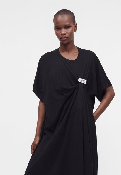 Femme aux cheveux courts portant une robe noire ample avec une étiquette rectangulaire blanche suspendue de la poitrine sur un fond uni.