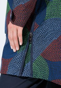 Ruka odpočívající na mnohobarevné bundě s velkým černým zipem a vzorem modrých, červených, zelených a světle modrých tečkovaných čar.