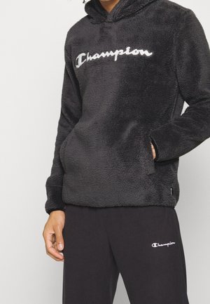 Personne portant un sweat à capuche noir duveteux et un pantalon de survêtement noir, tous deux avec le logo blanc "Champion", une main dans la poche du sweat.