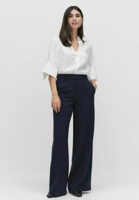 Chemise en lin blanc avec des manches retroussées, associée à un pantalon large bleu marine. Les deux vêtements ont une texture lisse et une coupe ajustée.