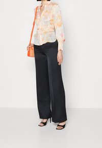 Femme portant un chemisier floral transparent, un pantalon large taille haute noir, des sandales à talons noires et portant un petit sac à bandoulière orange vif.