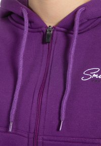 Lilla zip-up hættejakke lavet af blødt stof, med snørehætte, broderet logo og en metallisk lynlås lukning med matchende placket.