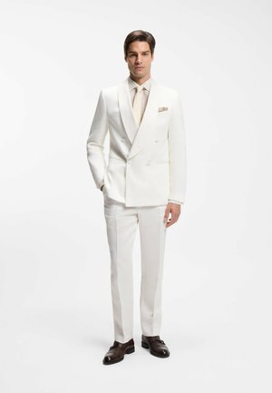 Man standing in a white double-breasted suit, cream shirt, tie, pocket square, and dark brown dress shoes against a white background.