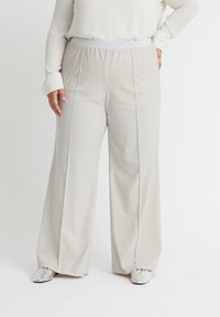 Beige weit geschnittene Hose aus weichem Stoff, mit einem elastischen Bund und vorderen Taschen, kombiniert mit silbernem Schuhwerk.