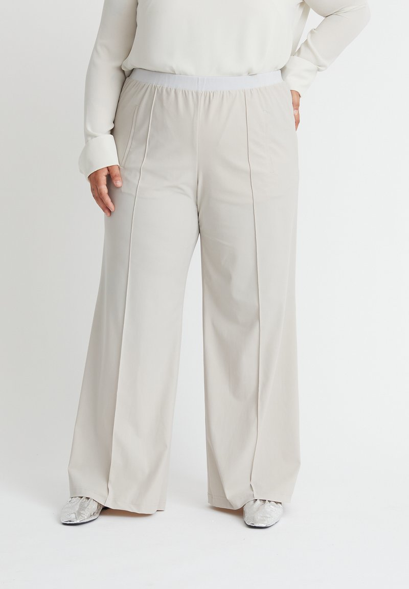 Beige weit geschnittene Hose aus weichem Stoff, mit einem elastischen Bund und vorderen Taschen, kombiniert mit silbernem Schuhwerk.