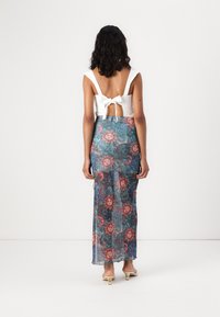 Witte, mouwloze top met strik aan de achterkant; lange, transparante rok met een kleurrijk bloemen- en paisleypatroon in blauwe, roze en groene tinten.