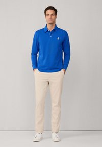 Blaues langärmliges Poloshirt mit Kragen und drei Knöpfen, kombiniert mit beigen Hosen und weißen Sneakers. Einfaches Design ohne Muster.