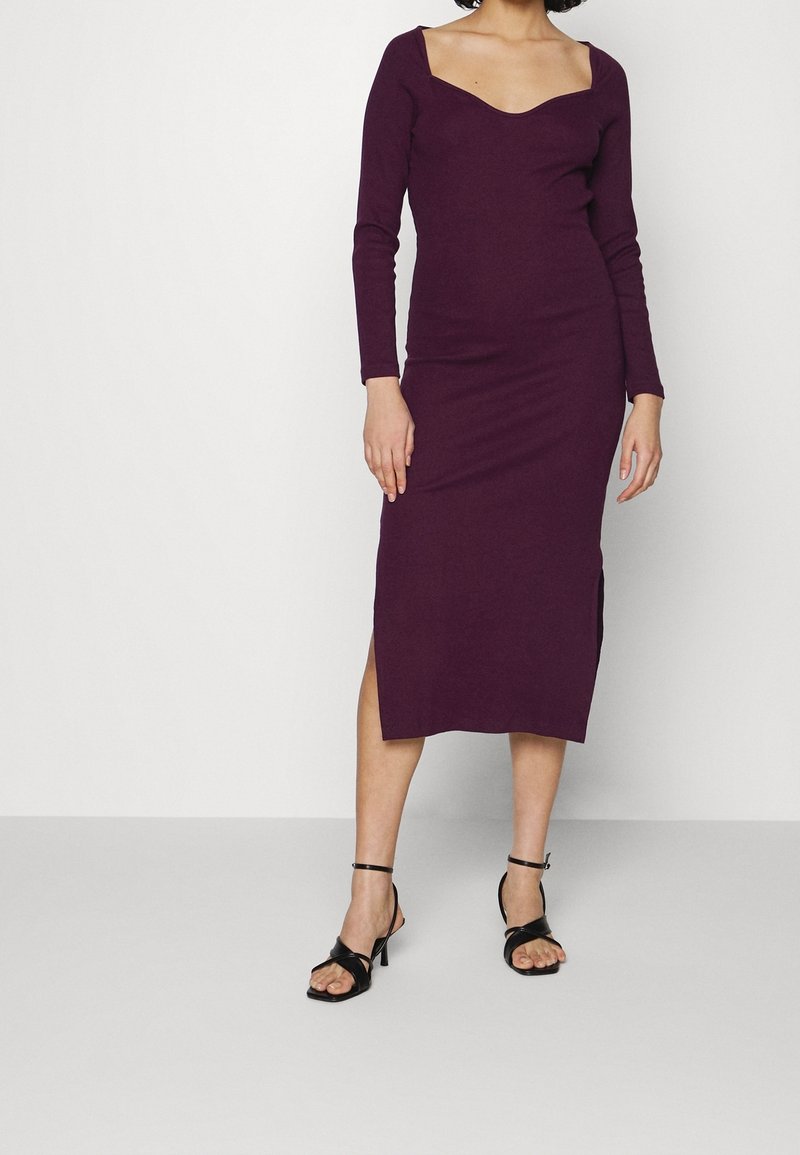 Femme portant une robe prune à manches longues, longueur midi, avec un décolleté carré et des sandales à talons hauts noires à brides, debout contre un mur blanc.