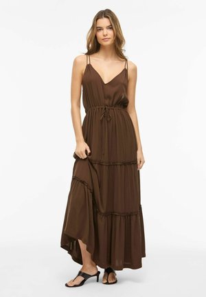 Ung kvinde i løs brun ærmeløs maxikjole og sorte sandaler med hæl, stående mod hvid baggrund.